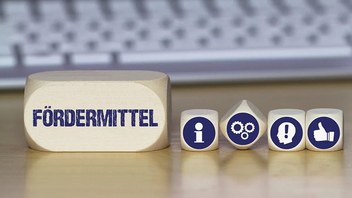 Ein Holzklotz mit der Aufschrift Fördermittel. Daneben liegen Würfel mit den Symbolen i (für Information), Zahnrädchen, ein Kopf mit einem Ausrufezeichen und ein Daumen-hoch.