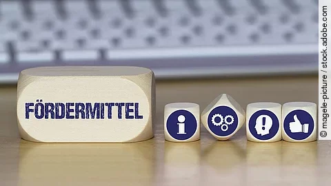 Ein Holzklotz mit der Aufschrift Fördermittel. Daneben liegen Würfel mit den Symbolen i (für Information), Zahnrädchen, ein Kopf mit einem Ausrufezeichen und ein Daumen-hoch.