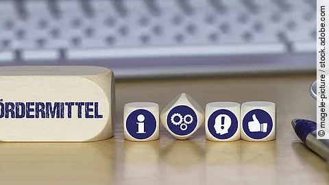 Ein Holzklotz mit der Aufschrift Fördermittel. Daneben liegen Würfel mit den Symbolen i (für Information), Zahnrädchen, ein Kopf mit einem Ausrufezeichen und ein Daumen-hoch.