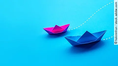 Zwei Papierboote in pink und blau fahren gemeinsam in eine Richtung.