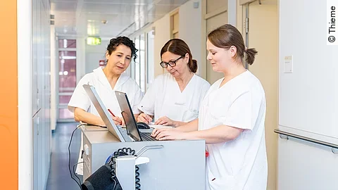 Drei Frauen in weißen Kasacks schauen in ein Notebook, was auf dem Stationswagen im Krankenhausflur steht.