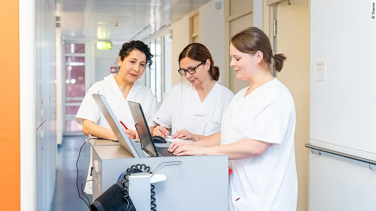 Drei Frauen in weißen Kasacks schauen in ein Notebook, was auf dem Stationswagen im Krankenhausflur steht.