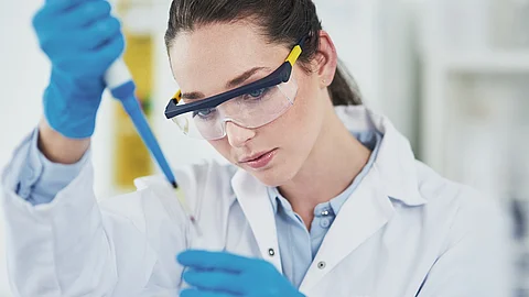 Eine Frau mit Schutzbrille und blauen Handschuhen hält eine Pipette in ein Reagenzglas 