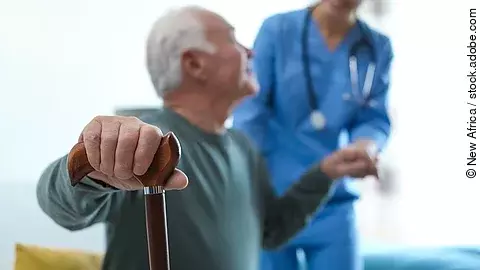 Eine Pflegekraft hilft einem sitzenden, älteren Patienten, der seine Hand am Gehstock hat