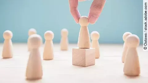 Mehrere Holzfiguren, die Personen darstellen. Eine der Figuren steht auf einem kleinen Würfel.