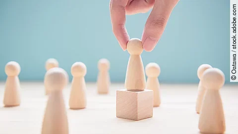 Mehrere Holzfiguren, die Personen darstellen. Eine der Figuren steht auf einem kleinen Würfel.