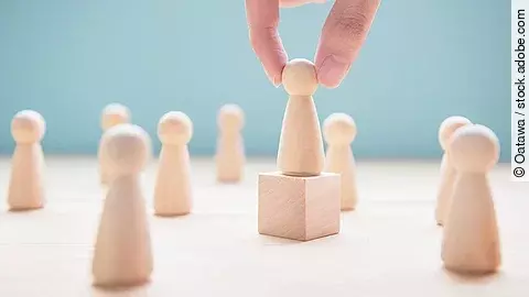 Mehrere Holzfiguren, die Personen darstellen. Eine der Figuren steht auf einem kleinen Würfel.