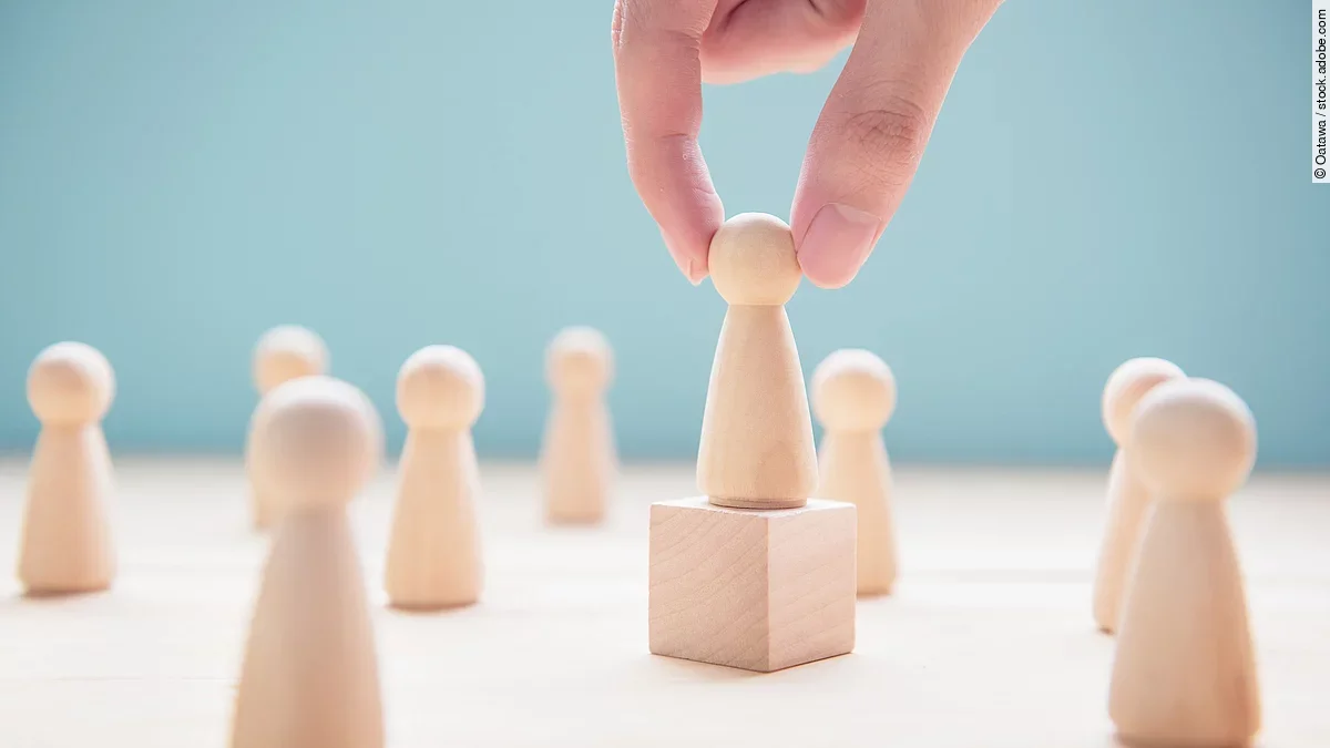 Mehrere Holzfiguren, die Personen darstellen. Eine der Figuren steht auf einem kleinen Würfel.