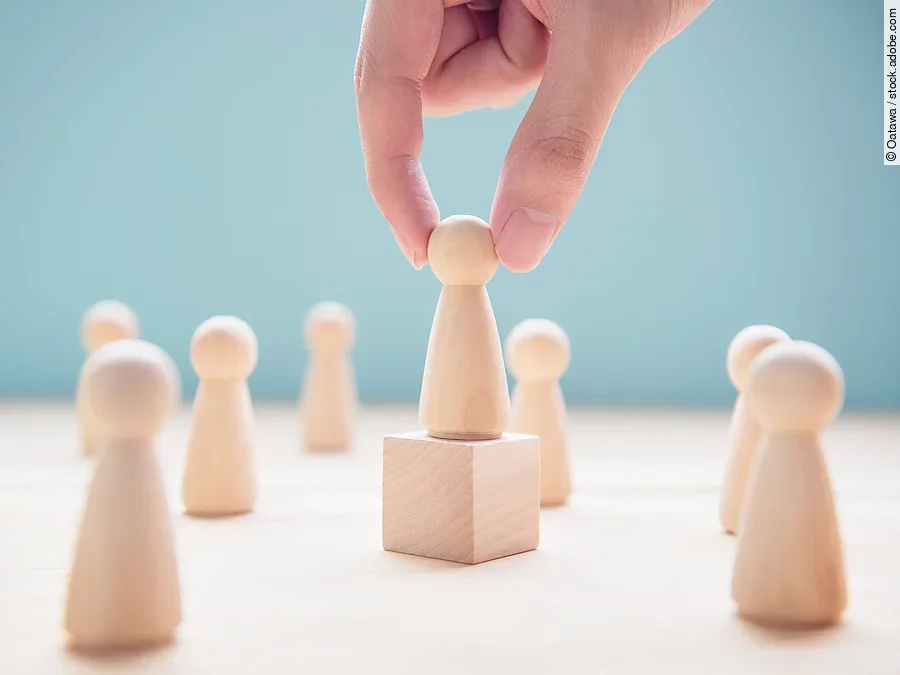 Mehrere Holzfiguren, die Personen darstellen. Eine der Figuren steht auf einem kleinen Würfel.