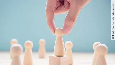 Mehrere Holzfiguren, die Personen darstellen. Eine der Figuren steht auf einem kleinen Würfel.