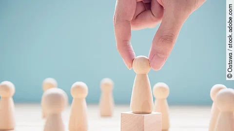 Mehrere Holzfiguren, die Personen darstellen. Eine der Figuren steht auf einem kleinen Würfel.