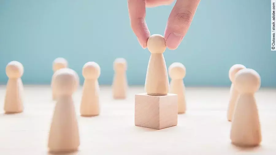 Mehrere Holzfiguren, die Personen darstellen. Eine der Figuren steht auf einem kleinen Würfel.