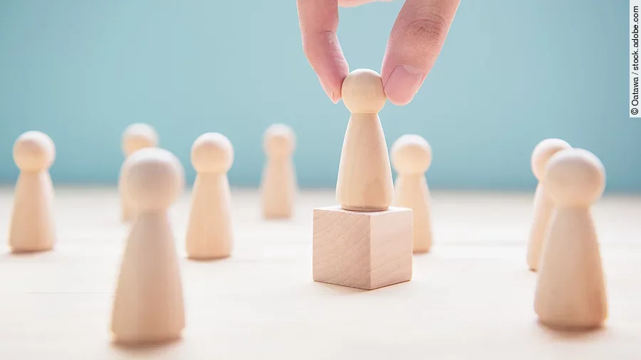 Mehrere Holzfiguren, die Personen darstellen. Eine der Figuren steht auf einem kleinen Würfel.