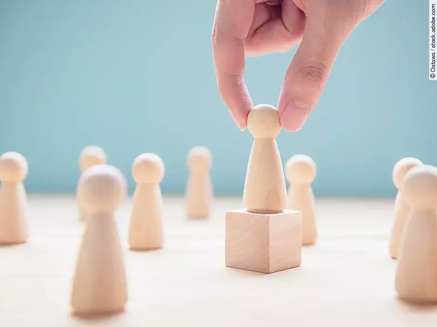 Mehrere Holzfiguren, die Personen darstellen. Eine der Figuren steht auf einem kleinen Würfel.