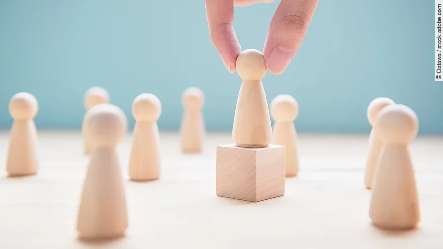 Mehrere Holzfiguren, die Personen darstellen. Eine der Figuren steht auf einem kleinen Würfel.
