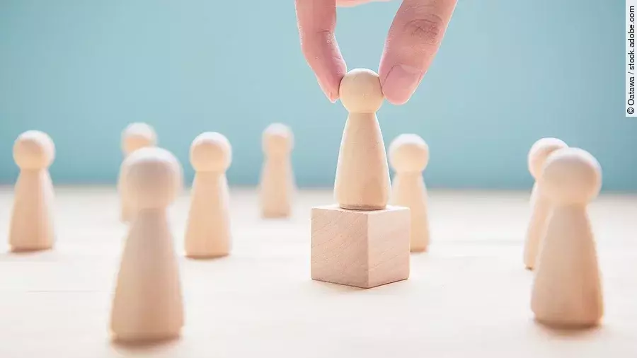 Mehrere Holzfiguren, die Personen darstellen. Eine der Figuren steht auf einem kleinen Würfel.