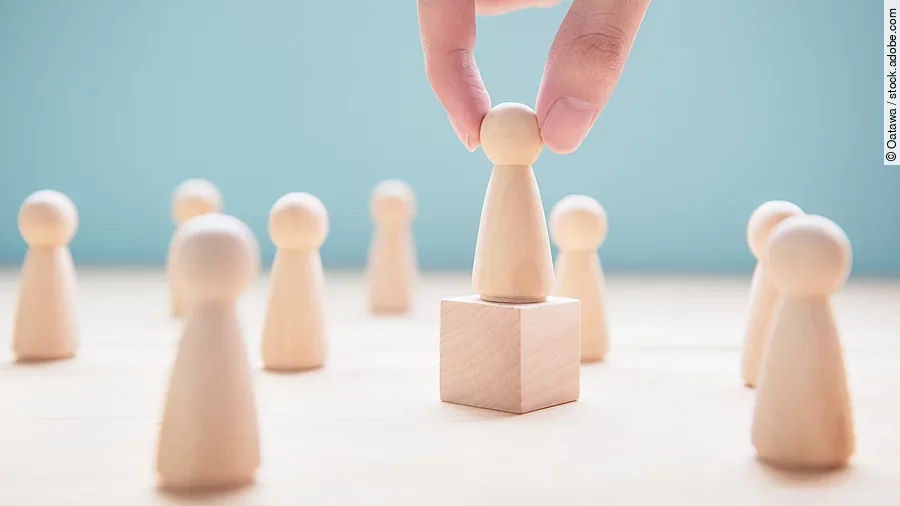 Mehrere Holzfiguren, die Personen darstellen. Eine der Figuren steht auf einem kleinen Würfel.