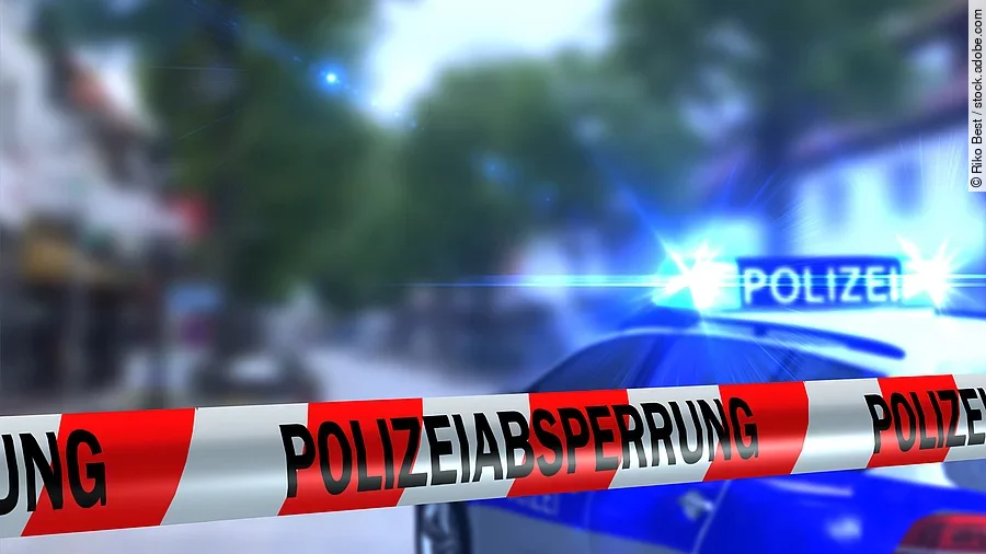 Polizeiabsperrung Ein rot-weißes Band mit der Beschriftung "Polizeiabsperrung". Im Hintergrund ist ein Polizeiauto zu sehen.