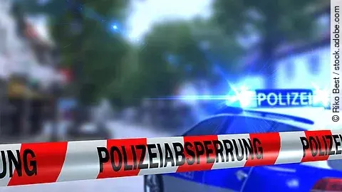 Ein rot-weißes Band mit der Beschriftung "Polizeiabsperrung". Im Hintergrund ist ein Polizeiauto zu sehen.