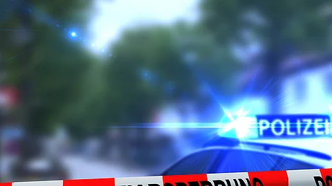 Ein rot-weißes Band mit der Beschriftung "Polizeiabsperrung". Im Hintergrund ist ein Polizeiauto zu sehen.