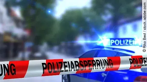 Ein rot-weißes Band mit der Beschriftung "Polizeiabsperrung". Im Hintergrund ist ein Polizeiauto zu sehen.