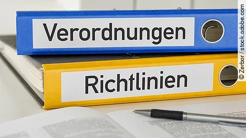 Verordnung Richtlinie Ein blauer Ordner mit der Beschriftung "Verordnungen" liegt auf einem gelben Ordner mit der Beschriftung "Richtlinien". Davor liegt ein grauer Kugelschreiber.