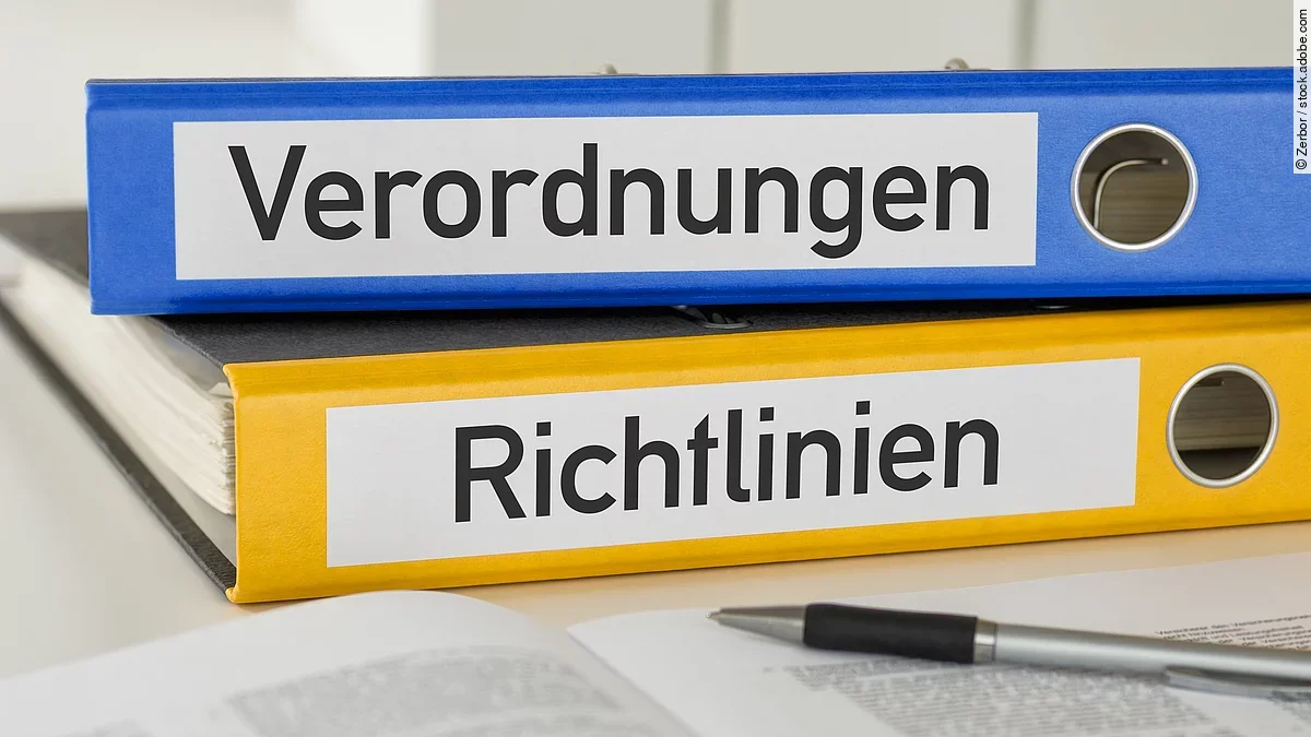 Ein blauer Ordner mit der Beschriftung "Verordnungen" liegt auf einem gelben Ordner mit der Beschriftung "Richtlinien". Davor liegt ein grauer Kugelschreiber.