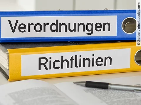 Ein blauer Ordner mit der Beschriftung "Verordnungen" liegt auf einem gelben Ordner mit der Beschriftung "Richtlinien". Davor liegt ein grauer Kugelschreiber.