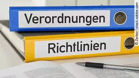 Ein blauer Ordner mit der Beschriftung "Verordnungen" liegt auf einem gelben Ordner mit der Beschriftung "Richtlinien". Davor liegt ein grauer Kugelschreiber.