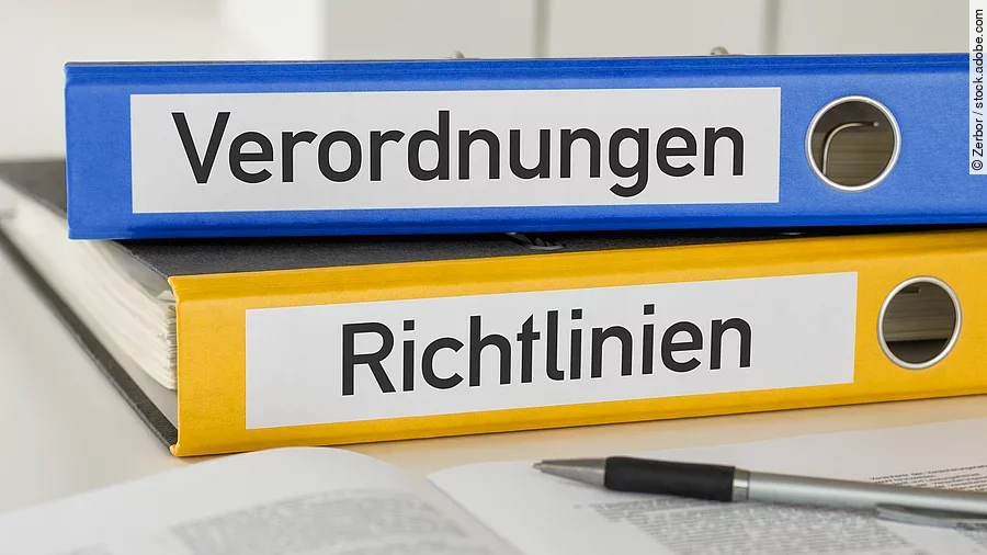 Verordnung Richtlinie Ein blauer Ordner mit der Beschriftung "Verordnungen" liegt auf einem gelben Ordner mit der Beschriftung "Richtlinien". Davor liegt ein grauer Kugelschreiber.