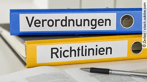 Ein blauer Ordner mit der Beschriftung "Verordnungen" liegt auf einem gelben Ordner mit der Beschriftung "Richtlinien". Davor liegt ein grauer Kugelschreiber.