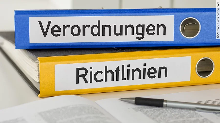 Ein blauer Ordner mit der Beschriftung "Verordnungen" liegt auf einem gelben Ordner mit der Beschriftung "Richtlinien". Davor liegt ein grauer Kugelschreiber.