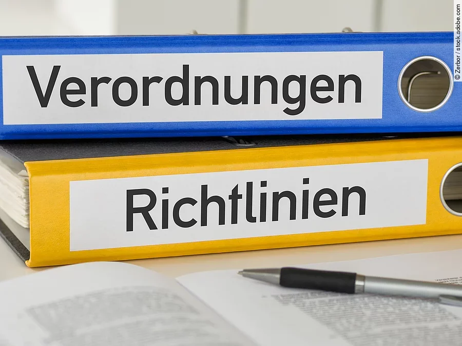 Ein blauer Ordner mit der Beschriftung "Verordnungen" liegt auf einem gelben Ordner mit der Beschriftung "Richtlinien". Davor liegt ein grauer Kugelschreiber.