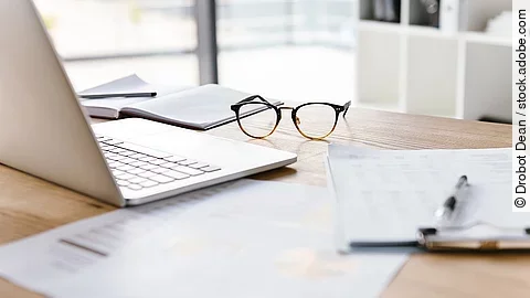 Arbeitsplatz Ein Arbeitsplatz in Form eines Holzschreibtisches. Darauf sind verschiedene Dokumente und eine Brille abgelegt. Ein Laptop steht aufgeklappt daneben.