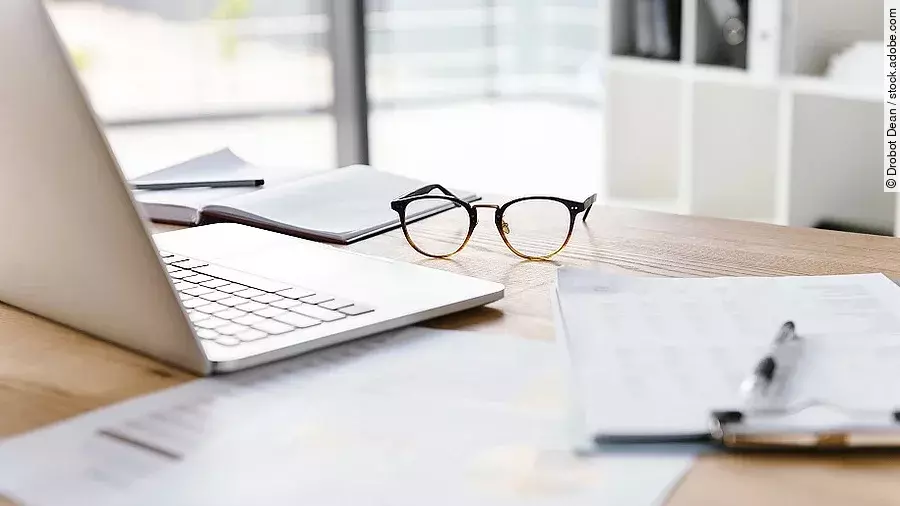 Ein Arbeitsplatz in Form eines Holzschreibtisches. Darauf sind verschiedene Dokumente und eine Brille abgelegt. Ein Laptop steht aufgeklappt daneben.