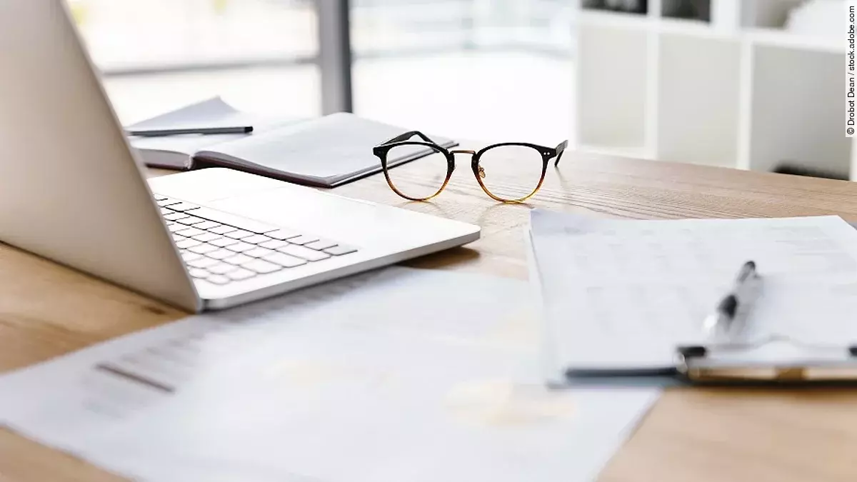 Arbeitsplatz Ein Arbeitsplatz in Form eines Holzschreibtisches. Darauf sind verschiedene Dokumente und eine Brille abgelegt. Ein Laptop steht aufgeklappt daneben.