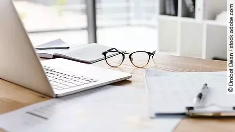 Arbeitsplatz Ein Arbeitsplatz in Form eines Holzschreibtisches. Darauf sind verschiedene Dokumente und eine Brille abgelegt. Ein Laptop steht aufgeklappt daneben.