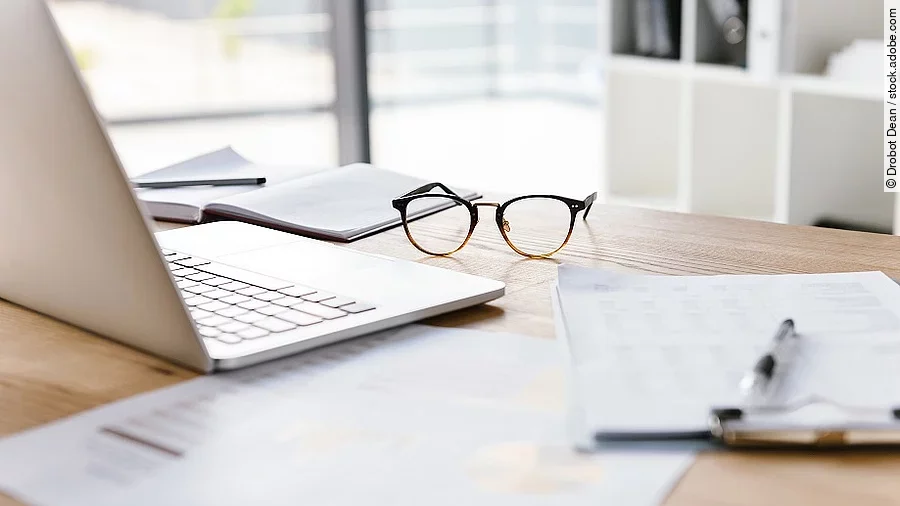 Ein Arbeitsplatz in Form eines Holzschreibtisches. Darauf sind verschiedene Dokumente und eine Brille abgelegt. Ein Laptop steht aufgeklappt daneben.