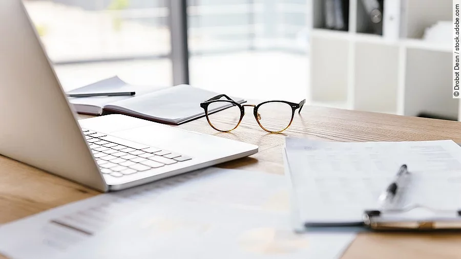 Ein Arbeitsplatz in Form eines Holzschreibtisches. Darauf sind verschiedene Dokumente und eine Brille abgelegt. Ein Laptop steht aufgeklappt daneben.