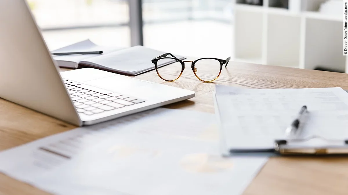 Arbeitsplatz Ein Arbeitsplatz in Form eines Holzschreibtisches. Darauf sind verschiedene Dokumente und eine Brille abgelegt. Ein Laptop steht aufgeklappt daneben.