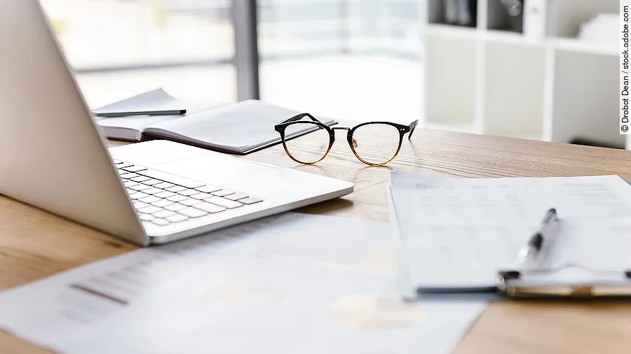Ein Arbeitsplatz in Form eines Holzschreibtisches. Darauf sind verschiedene Dokumente und eine Brille abgelegt. Ein Laptop steht aufgeklappt daneben.