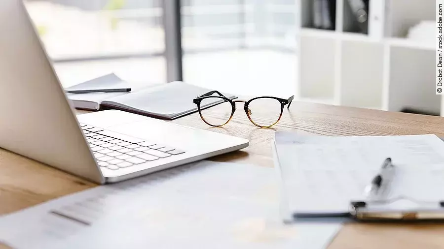 Arbeitsplatz Ein Arbeitsplatz in Form eines Holzschreibtisches. Darauf sind verschiedene Dokumente und eine Brille abgelegt. Ein Laptop steht aufgeklappt daneben.