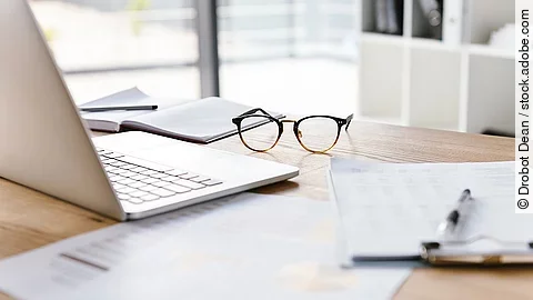 Ein Arbeitsplatz in Form eines Holzschreibtisches. Darauf sind verschiedene Dokumente und eine Brille abgelegt. Ein Laptop steht aufgeklappt daneben.