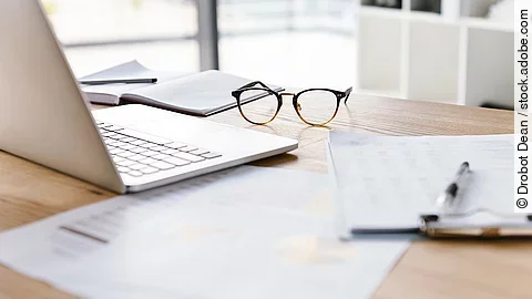 Arbeitsplatz Ein Arbeitsplatz in Form eines Holzschreibtisches. Darauf sind verschiedene Dokumente und eine Brille abgelegt. Ein Laptop steht aufgeklappt daneben.