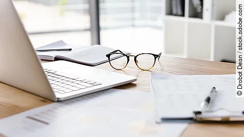 Arbeitsplatz Ein Arbeitsplatz in Form eines Holzschreibtisches. Darauf sind verschiedene Dokumente und eine Brille abgelegt. Ein Laptop steht aufgeklappt daneben.