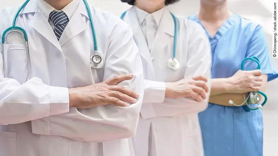 Drei Personen stehen nebeneinander. Links ein Arzt in weißem Kittel, in der Mitte eine Ärztin mit weißem Kittel, rechts eine Krankenschwester in blauem Kasack. Es sind jeweils nur die Torsi zu sehen. Alle drei haben ein Stethoskop.