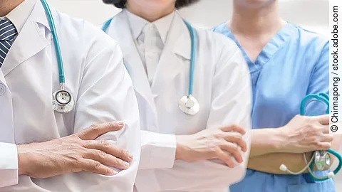 Drei Personen stehen nebeneinander. Links ein Arzt in weißem Kittel, in der Mitte eine Ärztin mit weißem Kittel, rechts eine Krankenschwester in blauem Kasack. Es sind jeweils nur die Torsi zu sehen. Alle drei haben ein Stethoskop.
