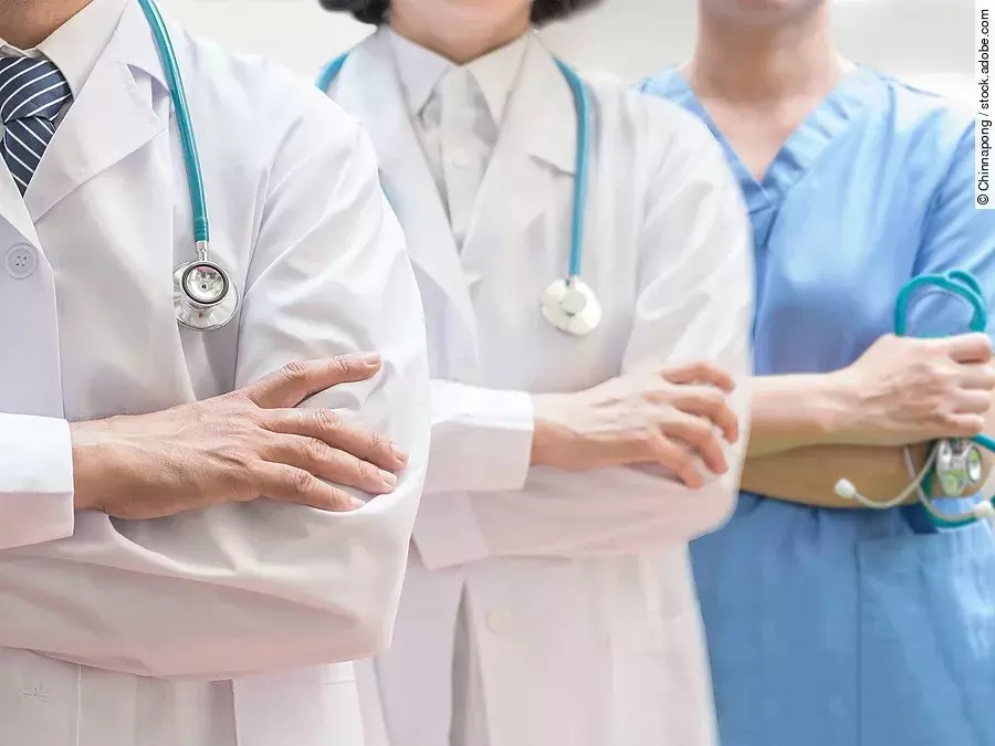 Drei Personen stehen nebeneinander. Links ein Arzt in weißem Kittel, in der Mitte eine Ärztin mit weißem Kittel, rechts eine Krankenschwester in blauem Kasack. Es sind jeweils nur die Torsi zu sehen. Alle drei haben ein Stethoskop.