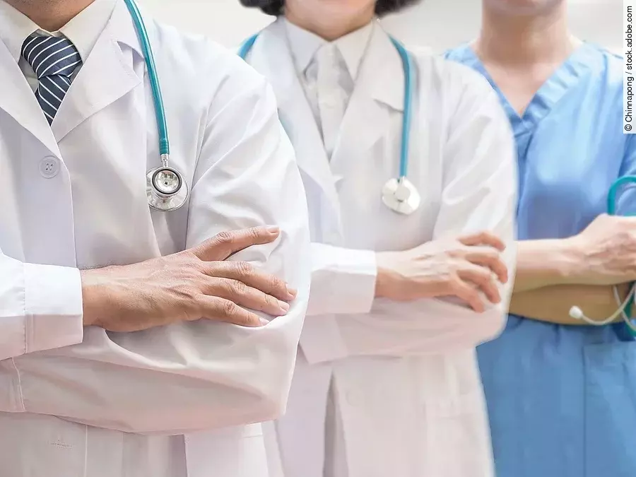 Drei Personen stehen nebeneinander. Links ein Arzt in weißem Kittel, in der Mitte eine Ärztin mit weißem Kittel, rechts eine Krankenschwester in blauem Kasack. Es sind jeweils nur die Torsi zu sehen. Alle drei haben ein Stethoskop.