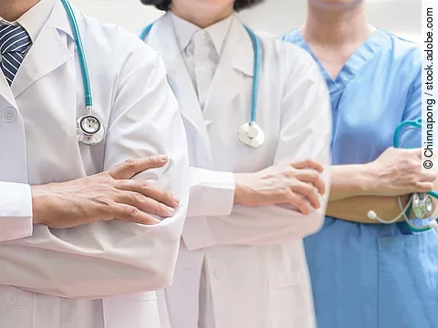 Drei Personen stehen nebeneinander. Links ein Arzt in weißem Kittel, in der Mitte eine Ärztin mit weißem Kittel, rechts eine Krankenschwester in blauem Kasack. Es sind jeweils nur die Torsi zu sehen. Alle drei haben ein Stethoskop.